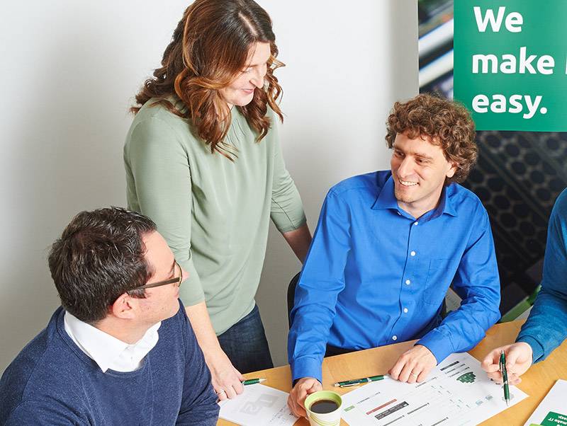 Eine Frau steht und zwei Männer sitzen an einem Besprechungstisch