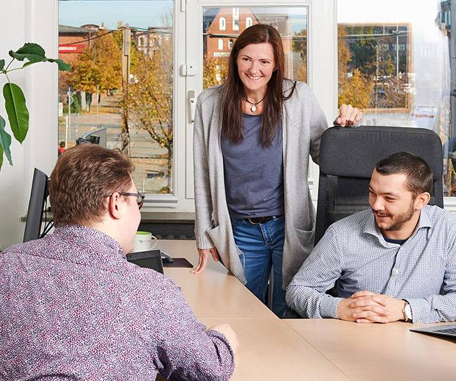 Frau steht am Büroisch mit zwei sitzenden Männern